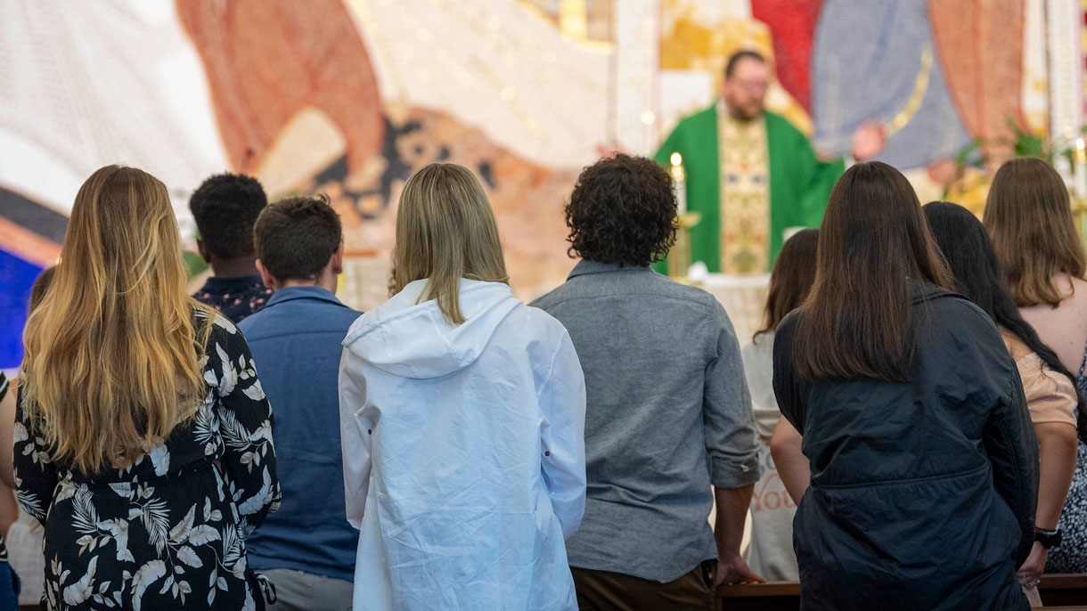 Students standing in chapel pews, priest at alter with hands raised