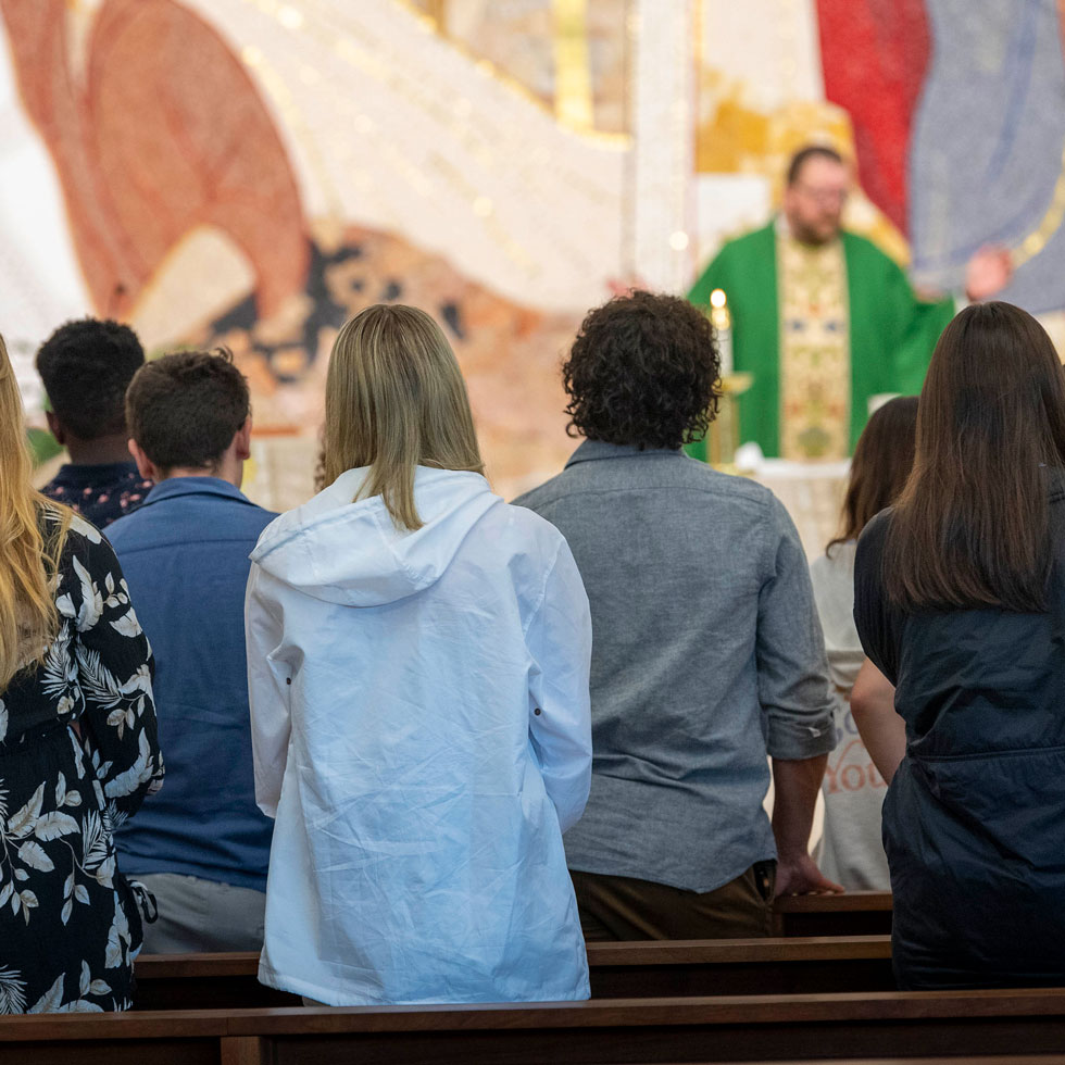 Students standing in chapel pews, priest at alter with hands raised