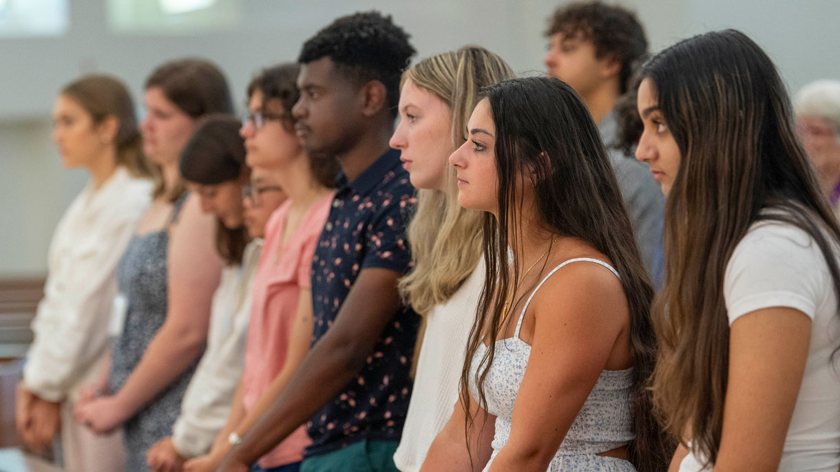 Students standing in chapel pews listening to the priest