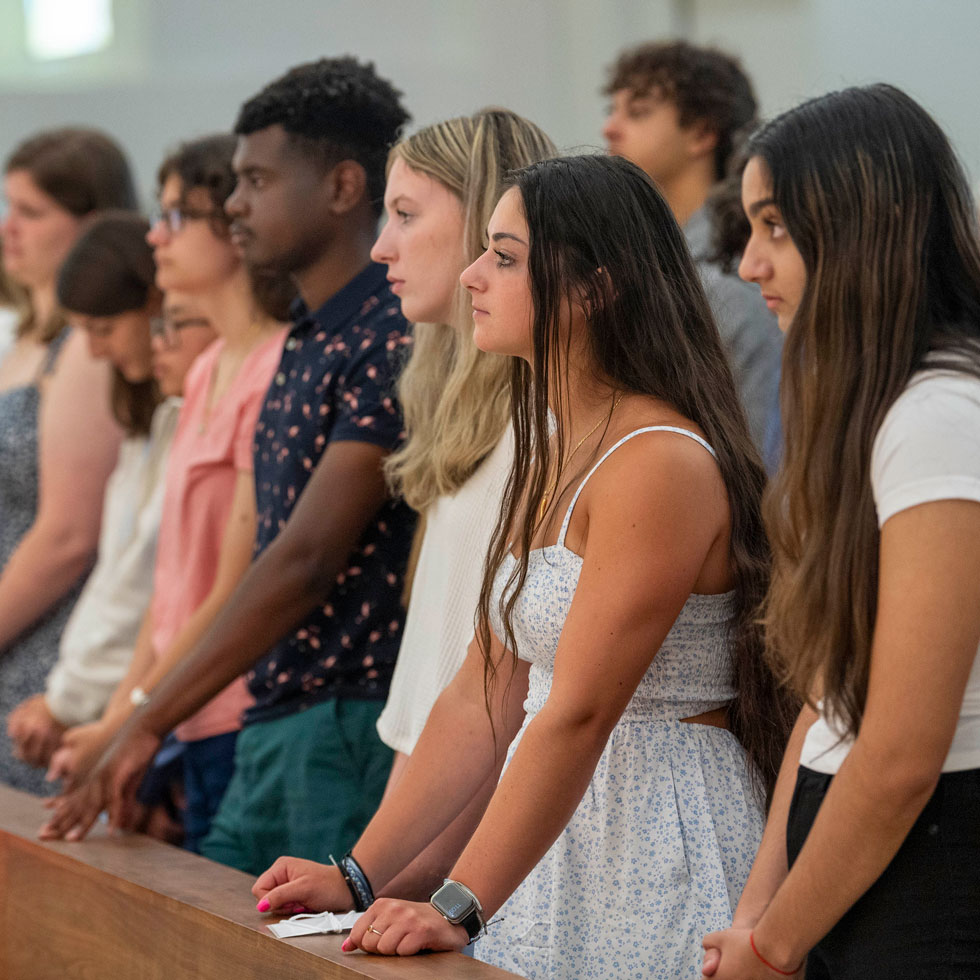 Students standing in chapel pews listening to the priest