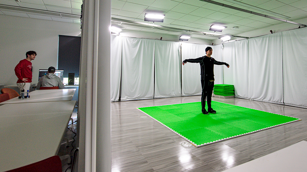 college student in a motion capture suit with classmates at a computer