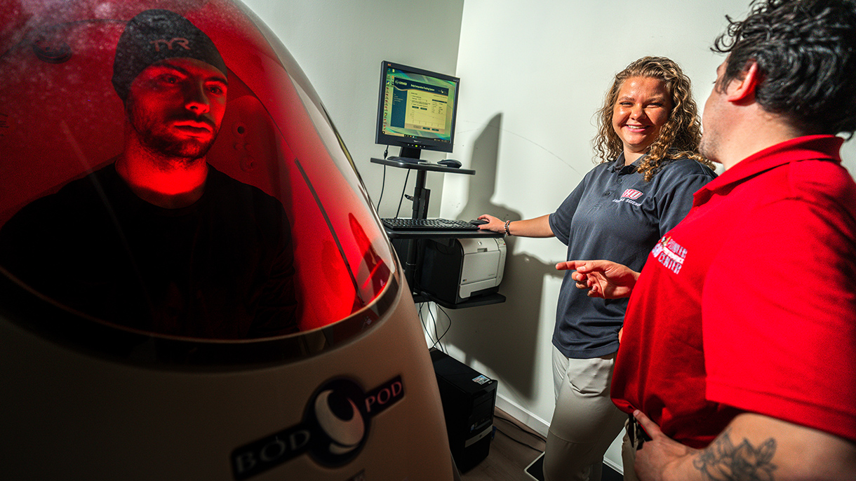 a person sitting in a bod pod while two exercise scientists monitor the test