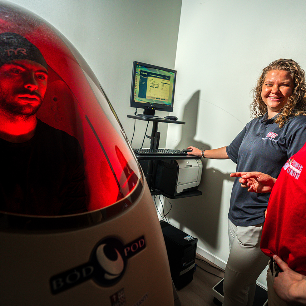 a person sitting in a bod pod while two exercise scientists monitor the test