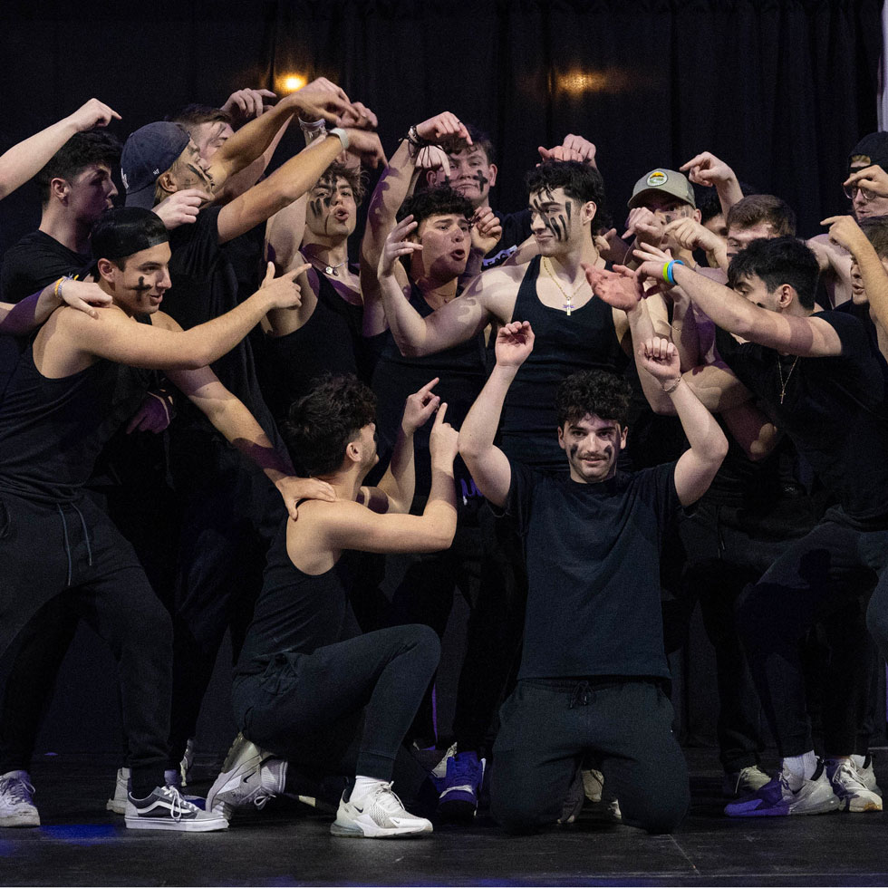 Members of Delta Tau Delta performing in Greek sing. All pointing towards one person and have black painted lines on their faces.