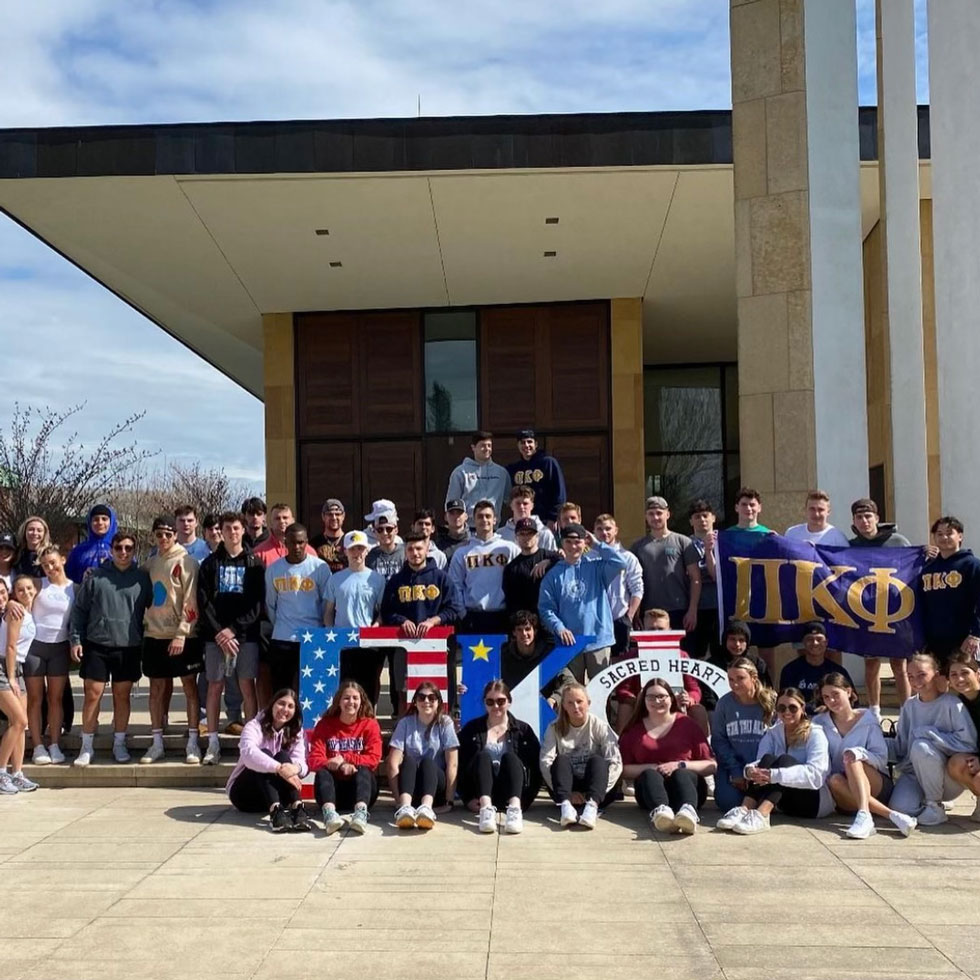 Members of Pi Kappa Phi as well as other Sacred Heart University peers stand in front of the chapel with their Greek Letters