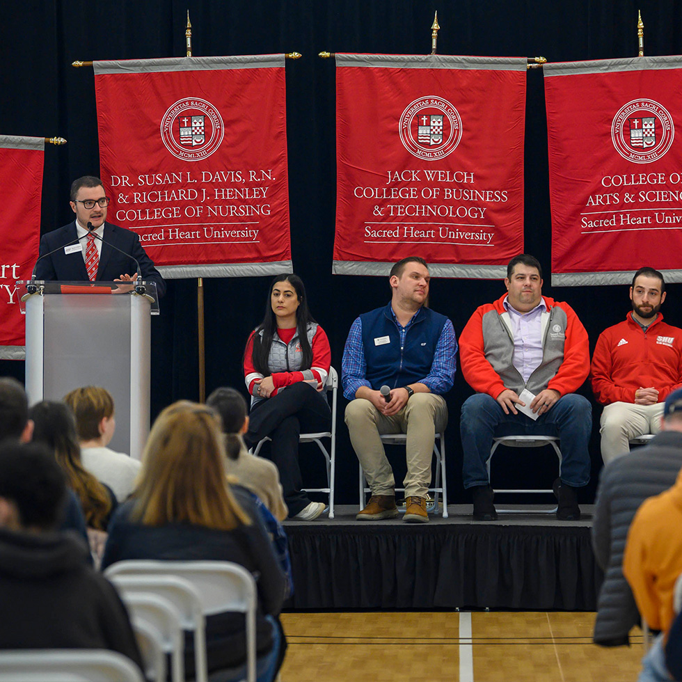 A man speaks at a podium with other speakers and banners behind him