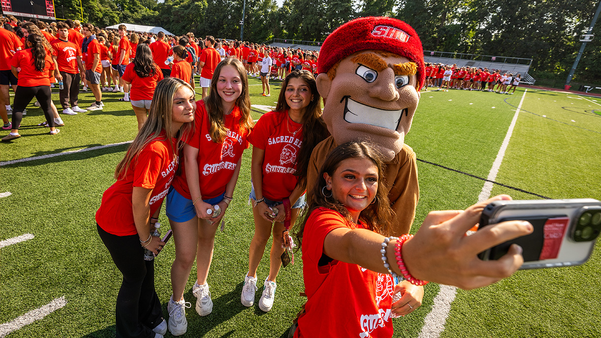 four people pose for a selfie on the SHU football field with mascot Big Red