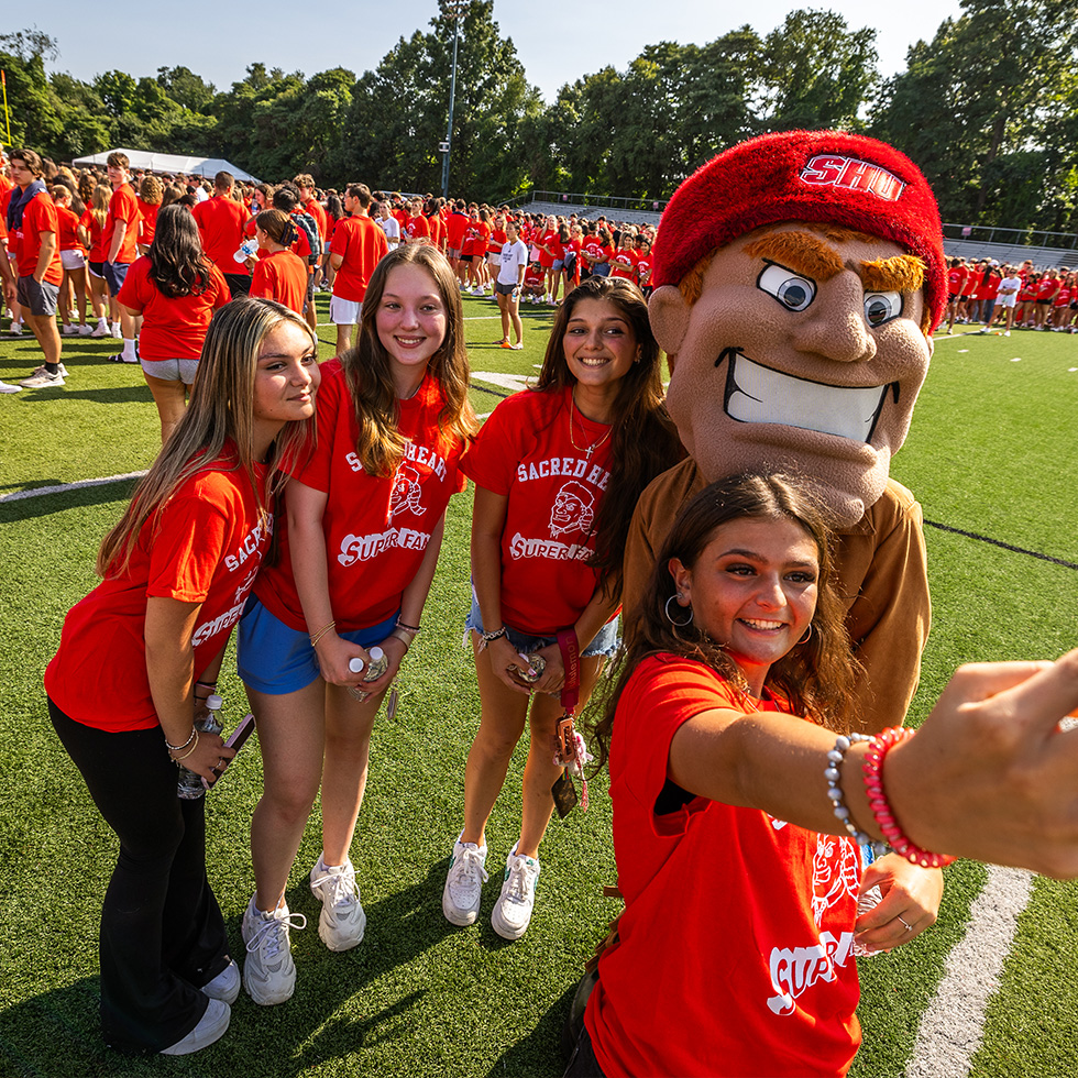 four people pose for a selfie on the SHU football field with mascot Big Red