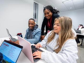 two college students working on laptops as their professor looks on