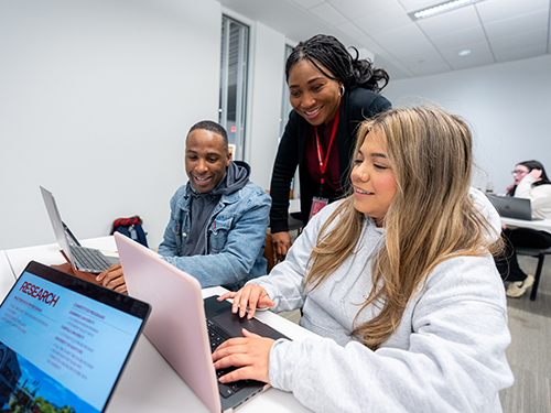 two college students working on laptops as their professor looks on