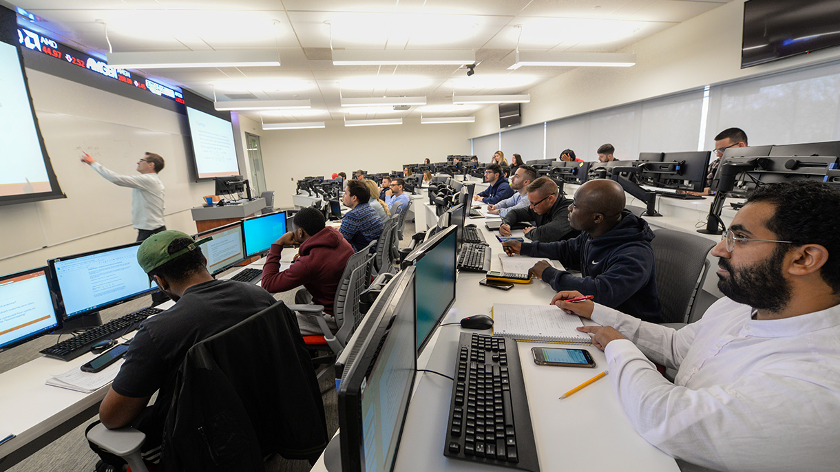 College students in a finance class watch the professor write on a whiteboard