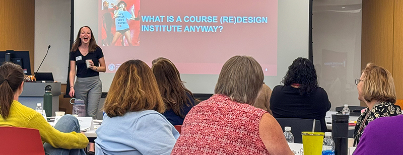 Person addressing a class with a slide on a screen behind them that says what is a course redesign institute anyway