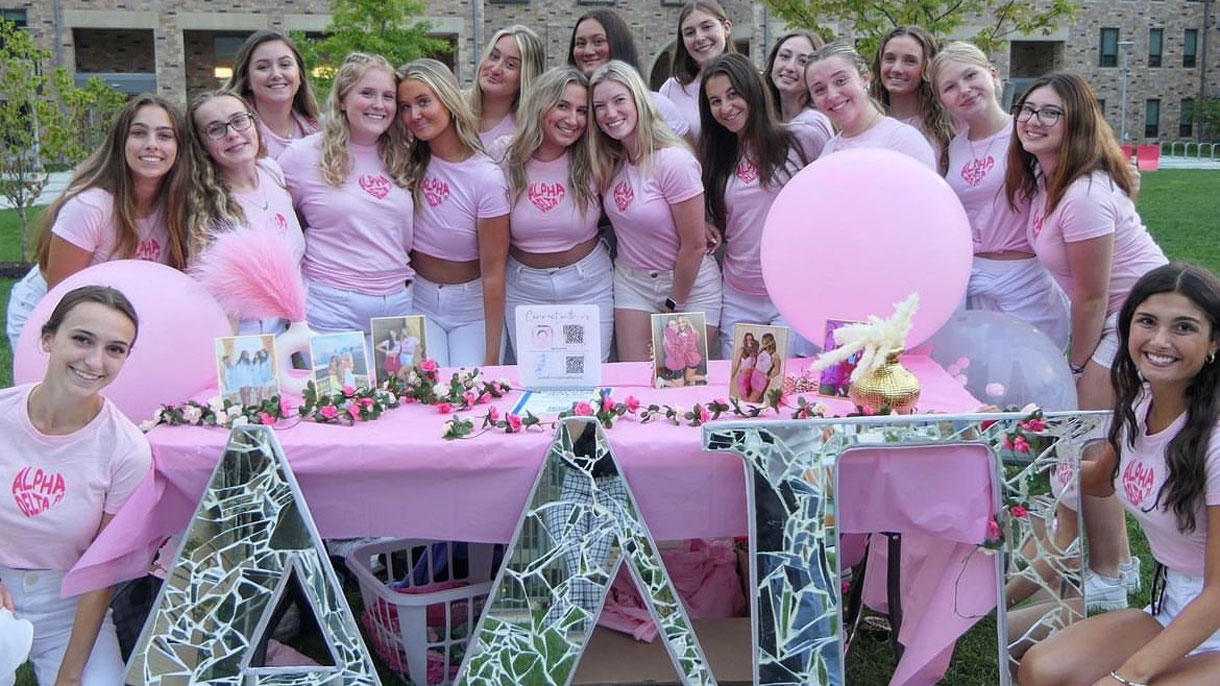 Members of Alpha Delta Pi posing for a photo dressed in pink t-shirts standing behind their sorority letters