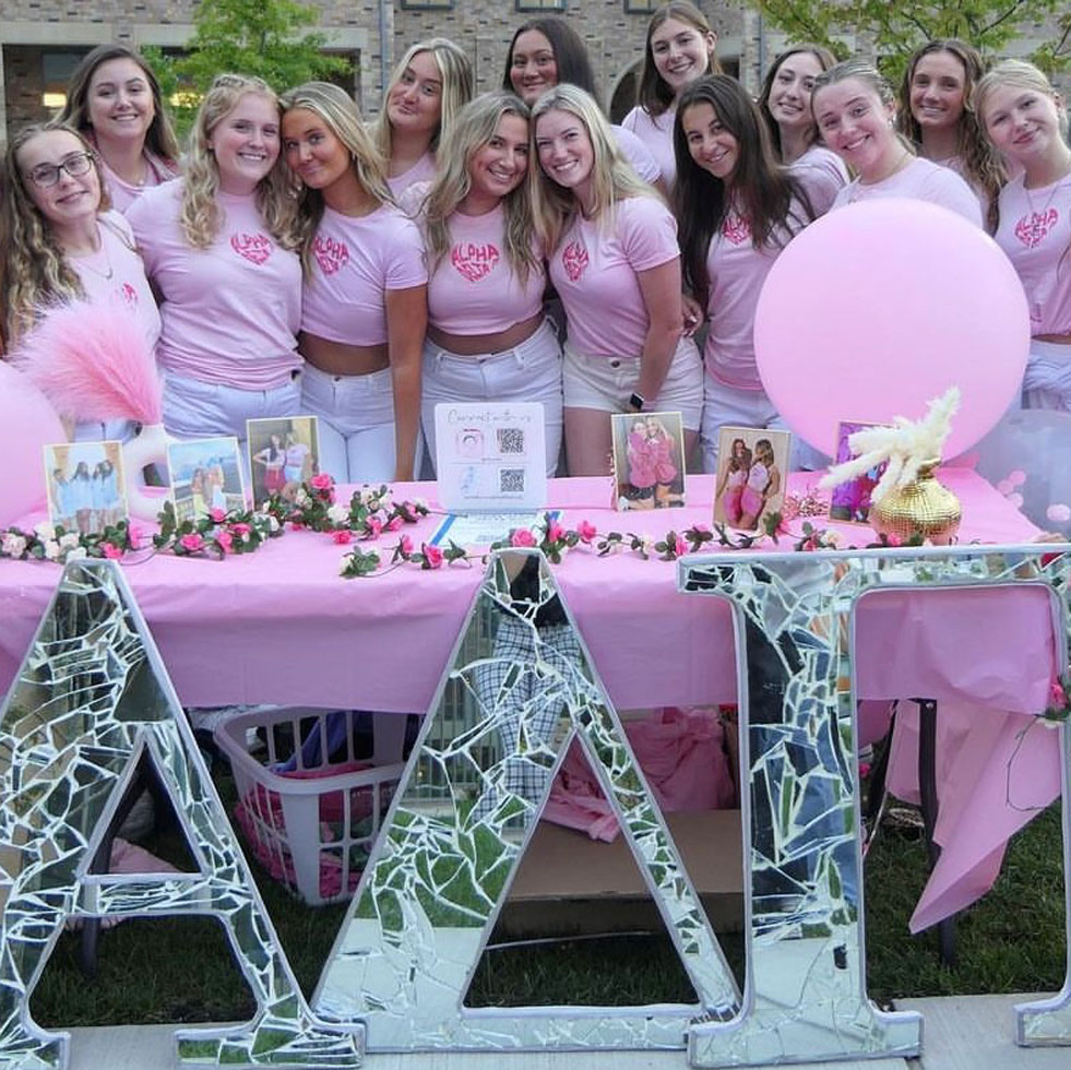 Members of Alpha Delta Pi posing for a photo dressed in pink t-shirts standing behind their sorority letters