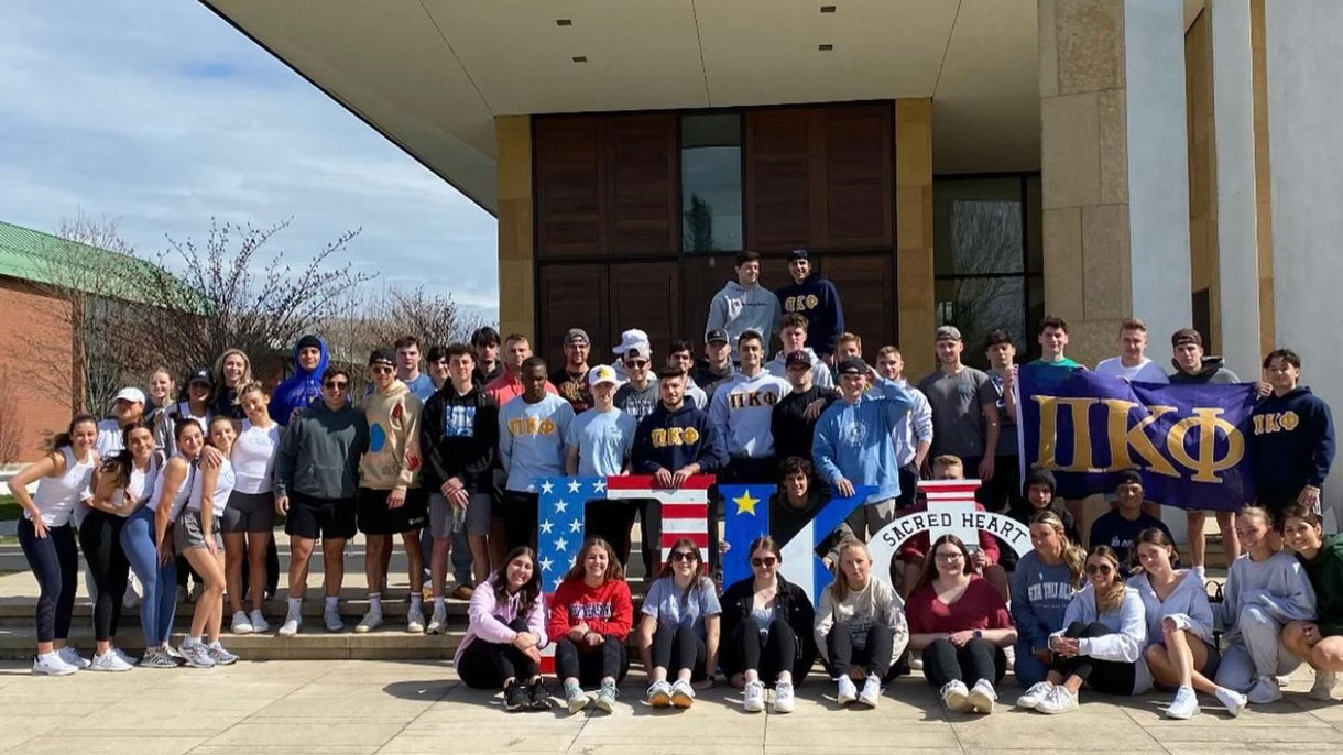 Members of Pi Kappa Phi as well as other Sacred Heart University peers stand in front of the chapel with their Greek Letters