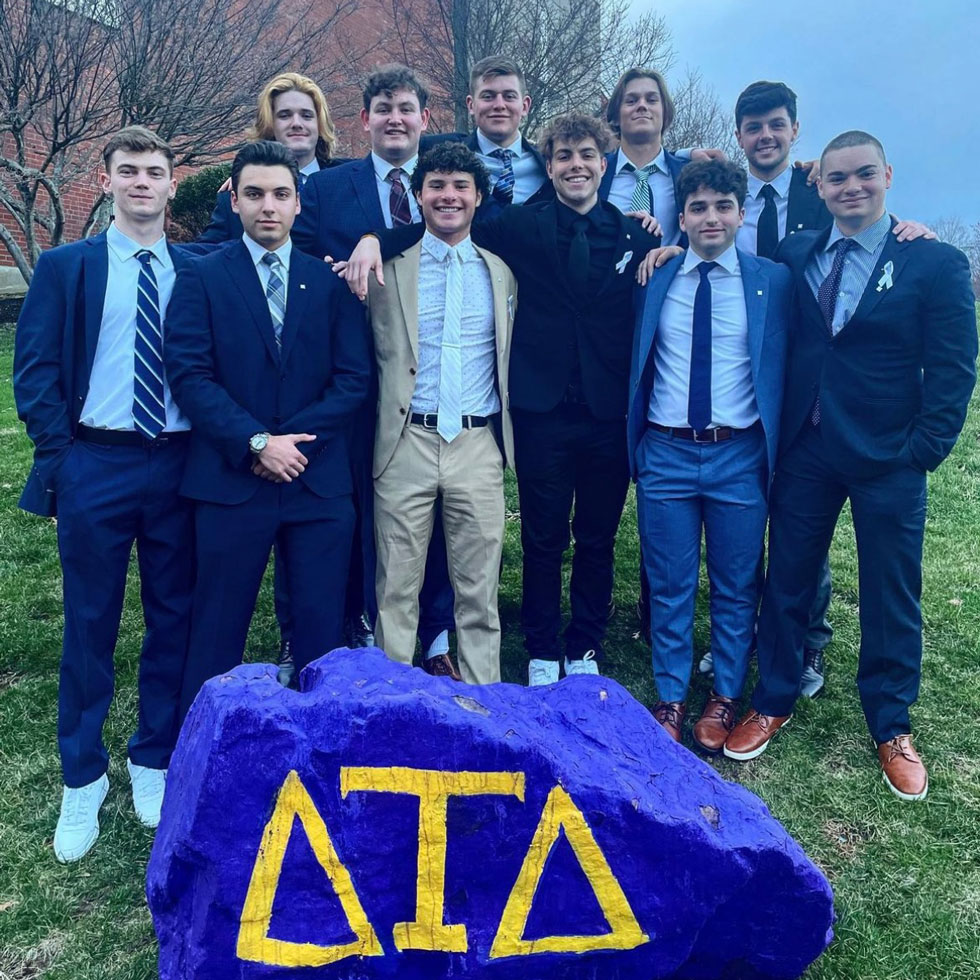 Members of Delta Tau Delta standing behind a rock with their fraternity letters painted on it.
