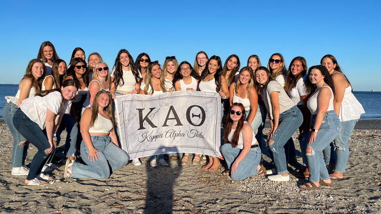 Members of Kappa Alpha Theta on the beach, wearing white shirts, holding a tapestry with their sorority letters on it