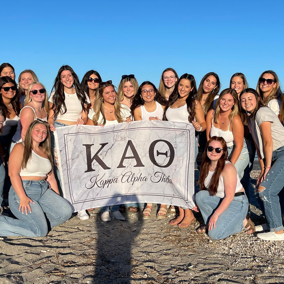 Members of Kappa Alpha Theta on the beach, wearing white shirts, holding a tapestry with their sorority letters on it