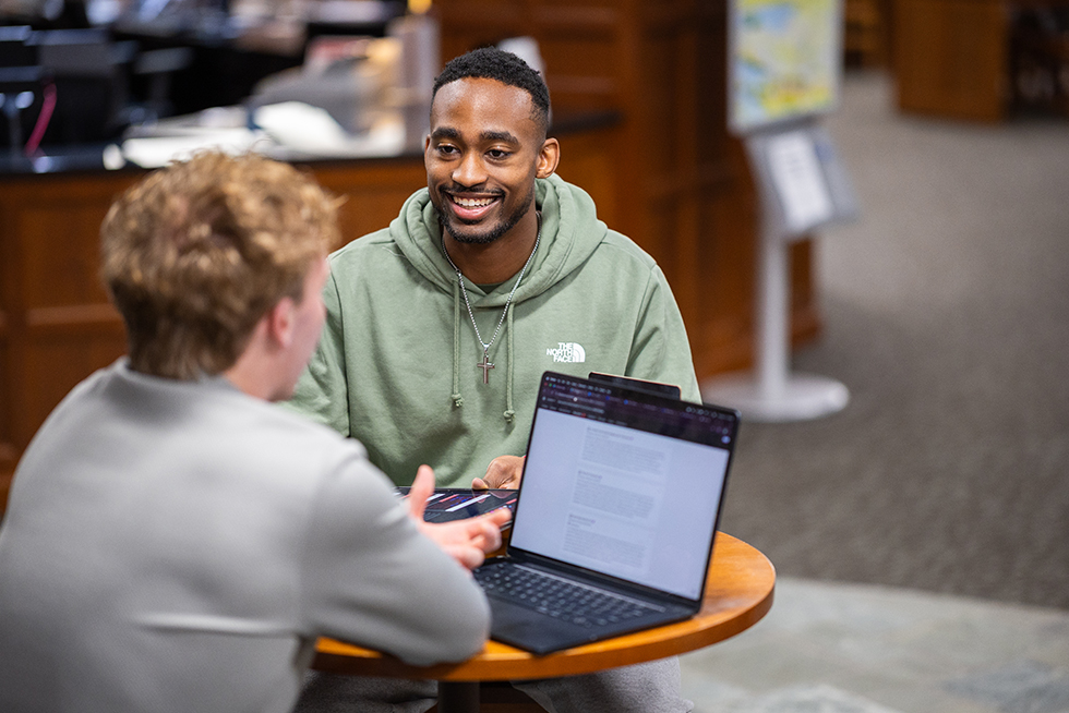 A smiling man talks with a friend who is looking at a laptop computer
