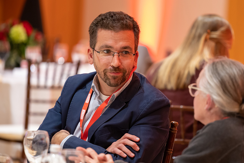 a man wearing glasses and a blazer in conversation with a woman