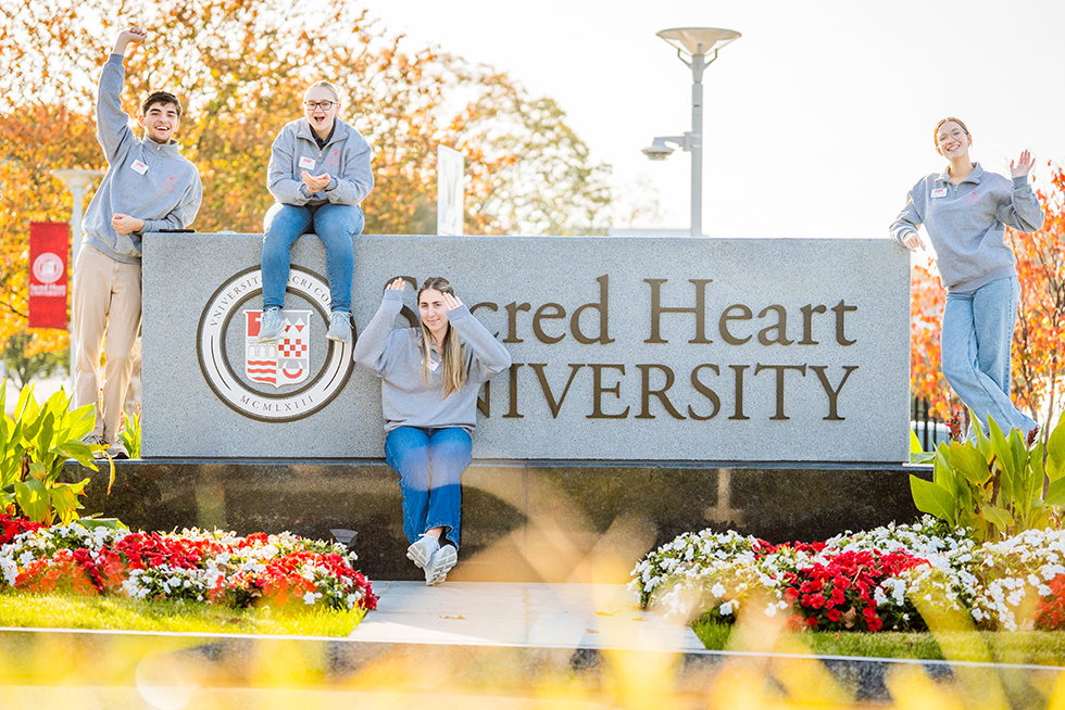 five people sitting and standing around a Sacred Heart University sign