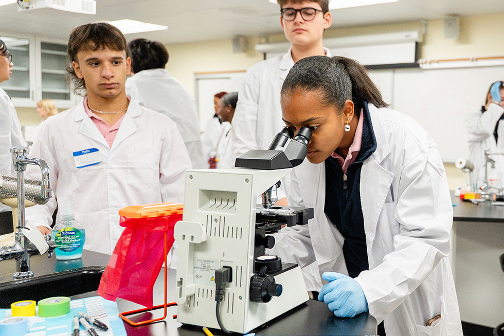 a biology student in a while lab coat looks into a microscope as two classmates look on