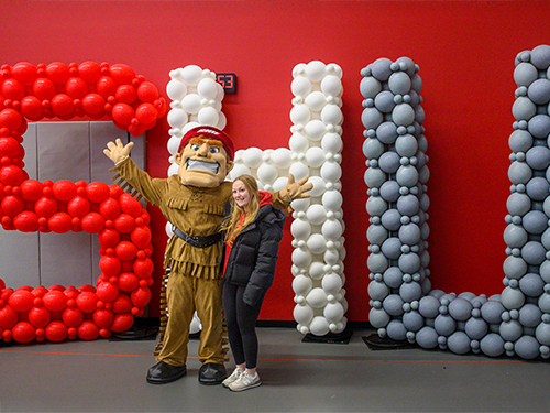 Big Red mascot poses with a student in front of balloons that spell out SHU