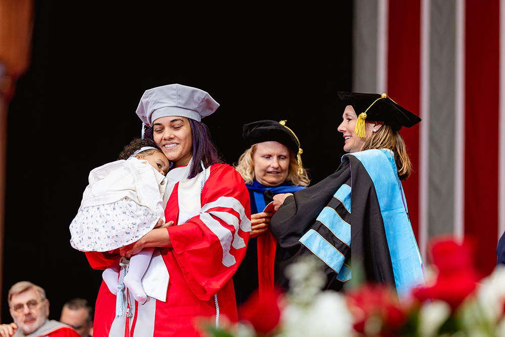 Graduate receives her hood while holding her baby
