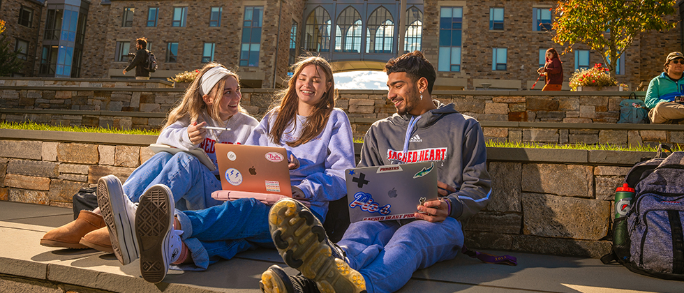 three college students sitting outside laughing together and working on their laptops