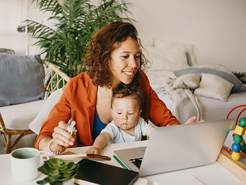 a smiling woman working on a laptop and holding a baby at the same time