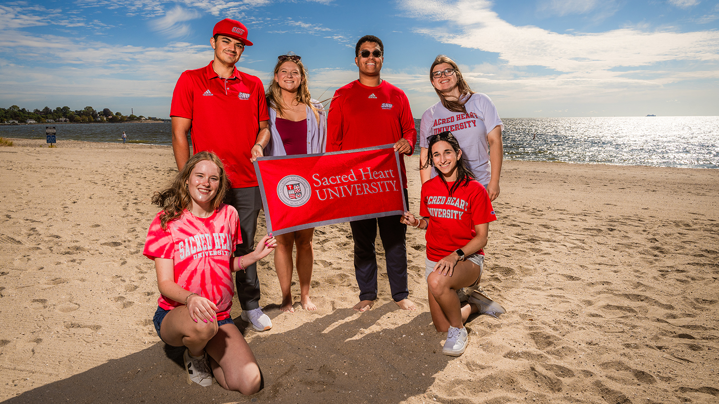 a group of college-age students posing for a photo on a beach with a Sacred Heart University banner