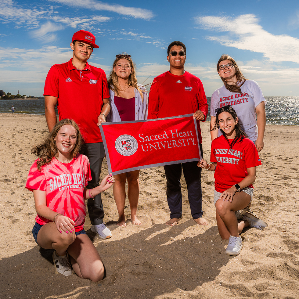 a group of college-age students posing for a photo on a beach with a Sacred Heart University banner