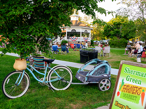 a town green showing a gazebo concert, a bicycle and a farmers market sign