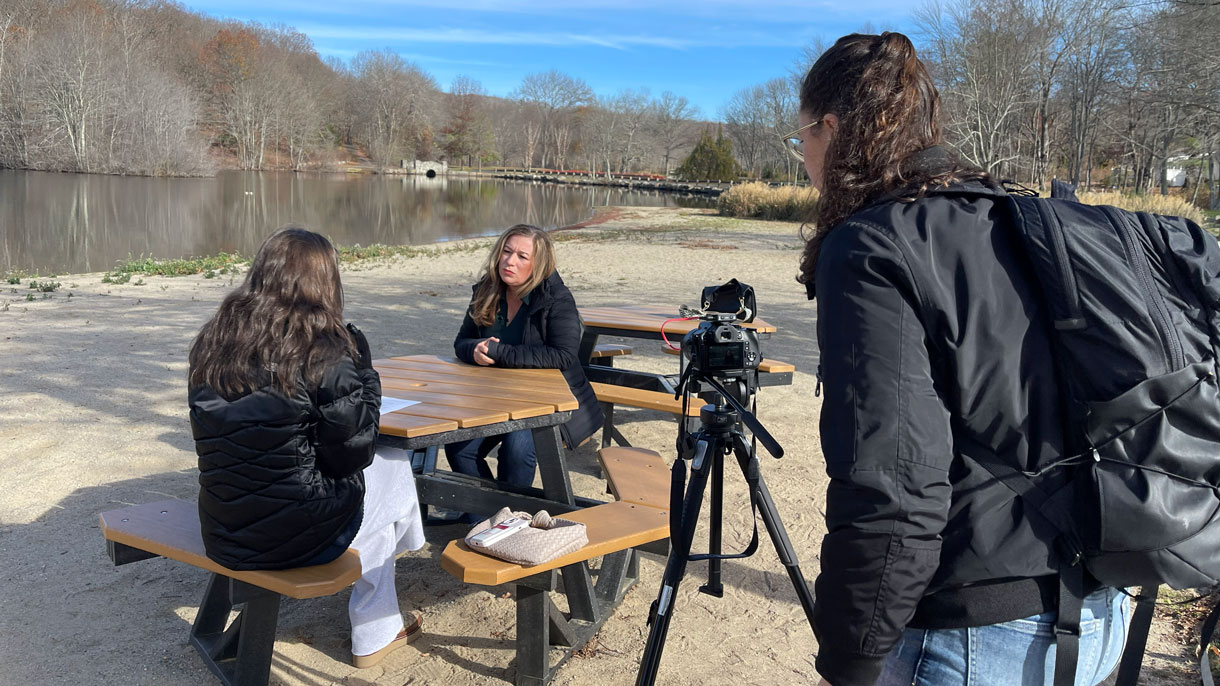 Student interviewing person next to lake while other student films