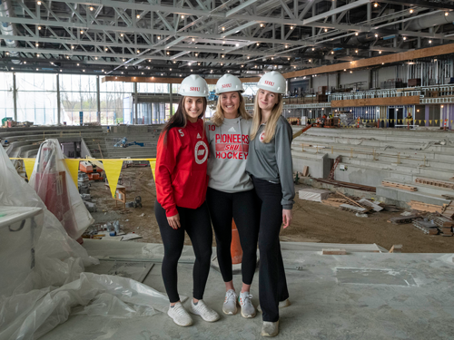 Three women in hard hats in a hockey arena being constructed