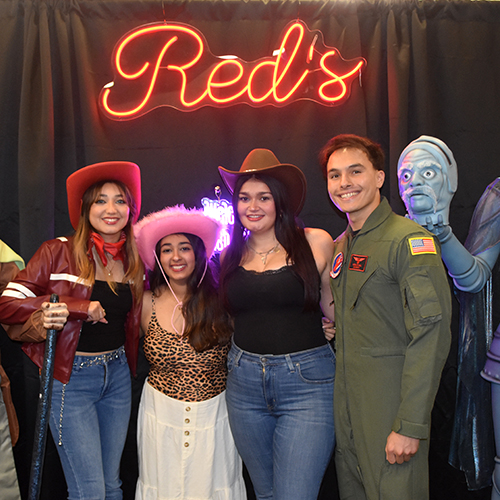 Group of people wearing Halloween costumes and standing in front of a sign that says Reds