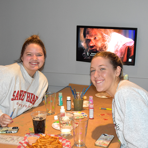 two people sitting at a table with painting supplies and drinks
