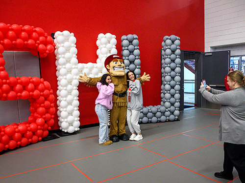 Big Red mascot posing for a photo with two people in front of a balloon display that spells out SHU