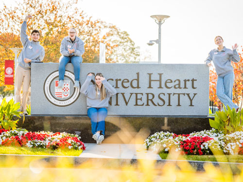 Students welcoming families in front of Sacred Heart University entrance sign